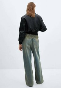 Femme aux cheveux roux mi-longs portant une veste bomber noire et un jean bleu délavé à jambes larges, debout devant un fond clair uni.