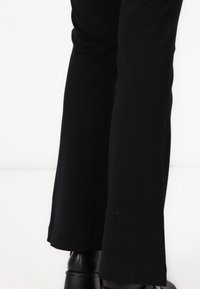 Pantalons évasés noirs en tissu lisse, présentant un design épuré avec une texture subtile au niveau de l'ourlet. Chaussures noires partiellement visibles.