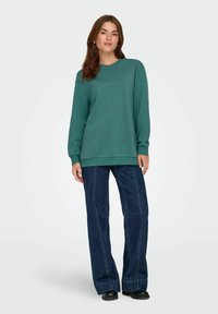 Felpa verde con maniche lunghe, collo rotondo e polsini a costine, abbinata a jeans in denim scuro a gamba larga. Tessuto dalla texture morbida.