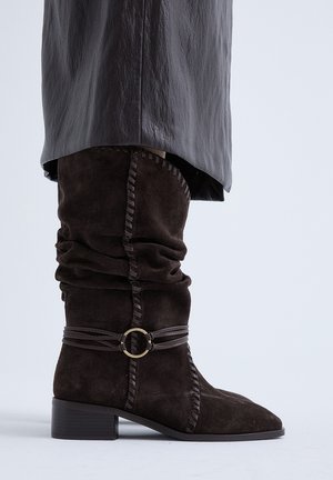 Bota de ante marrón oscuro hasta la mitad de la pantorrilla con diseño holgado, costuras decorativas cosidas, correa de cuero con anilla metálica, usada con falda de cuero oscuro.