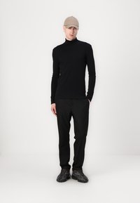 Suéter negro de manga larga y cuello alto, corte ajustado, combinado con pantalones negros y zapatillas oscuras. Una gorra beige completa el atuendo sobre un fondo liso.