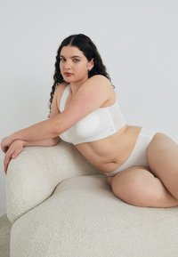 Hvit bralette og matchende undertøysett, med glatt stoff, minimalistisk design og bred skulderstropp. Bært av en modell på en teksturert sofa.