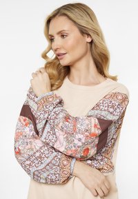 Ljust beige långärmad topp med mönstrade, flerfärgade blommönster på ärmarna. Har texturert tyg och sammetsfåll.