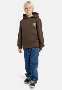 Hnědá mikina s kapucí a přední kapsou a s logem, v kombinaci s modrými džínami. Celkový vzhled doplňují tenisky s bílými podrážkami.