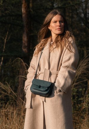 Green leather crossbody bag with a flap closure and stitched detailing, worn over a beige wool coat in a natural outdoor setting.