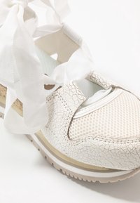 Närbild av vit sneakers med sprucken läderstruktur, mesh tyg tå, beige sula och knutna breda tygskosnören på en vit bakgrund.