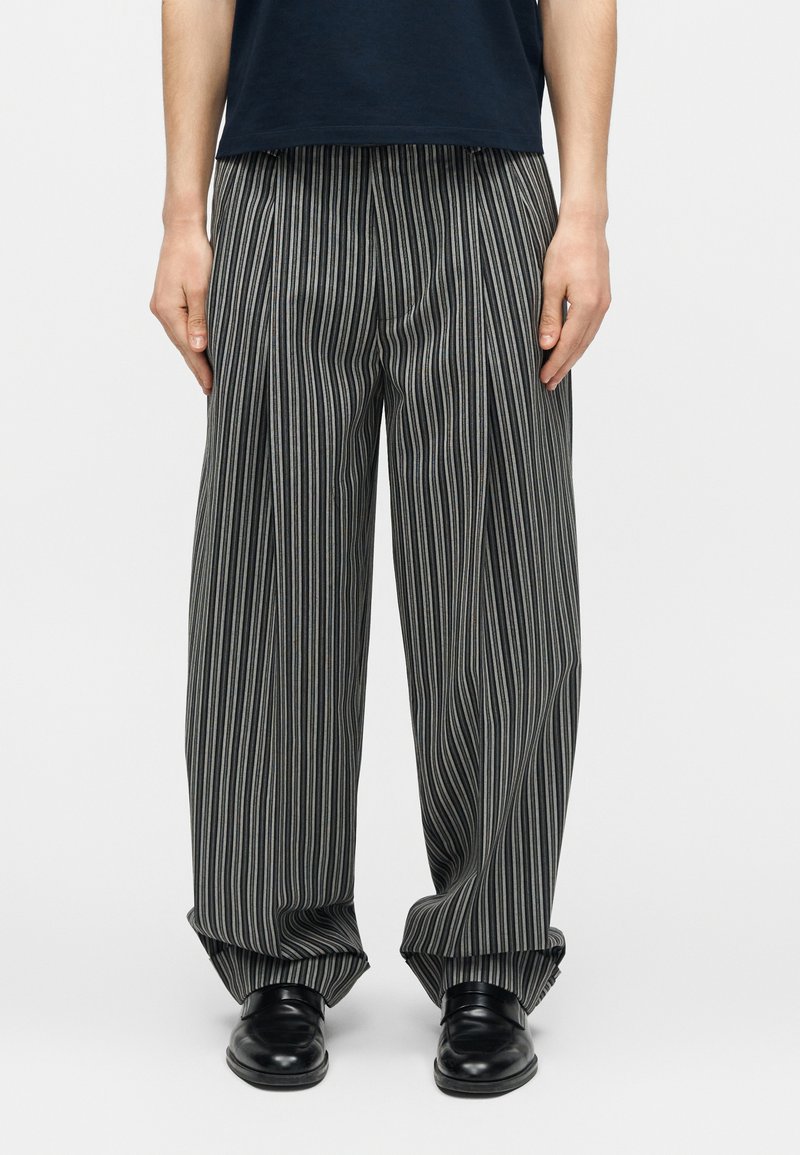 Persona che indossa pantaloni larghi a righe verticali nere e grigie con scarpe di pelle nere, in piedi contro uno sfondo semplice.