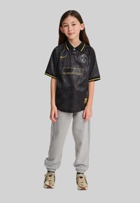 Svart polotröja med gula accenter, med ett mönstrat mönster och logotyp. Sammanfogad med grå sweatpants och gyllene sneakers.