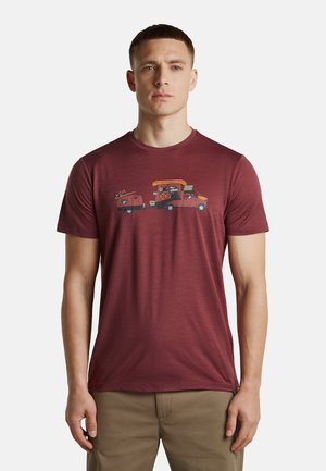 Junger Mann mit kurzen Haaren, der ein weinrotes T-Shirt mit einem Motiv eines Autos trägt, das einen kleinen Wohnwagen und Anhänger zieht.