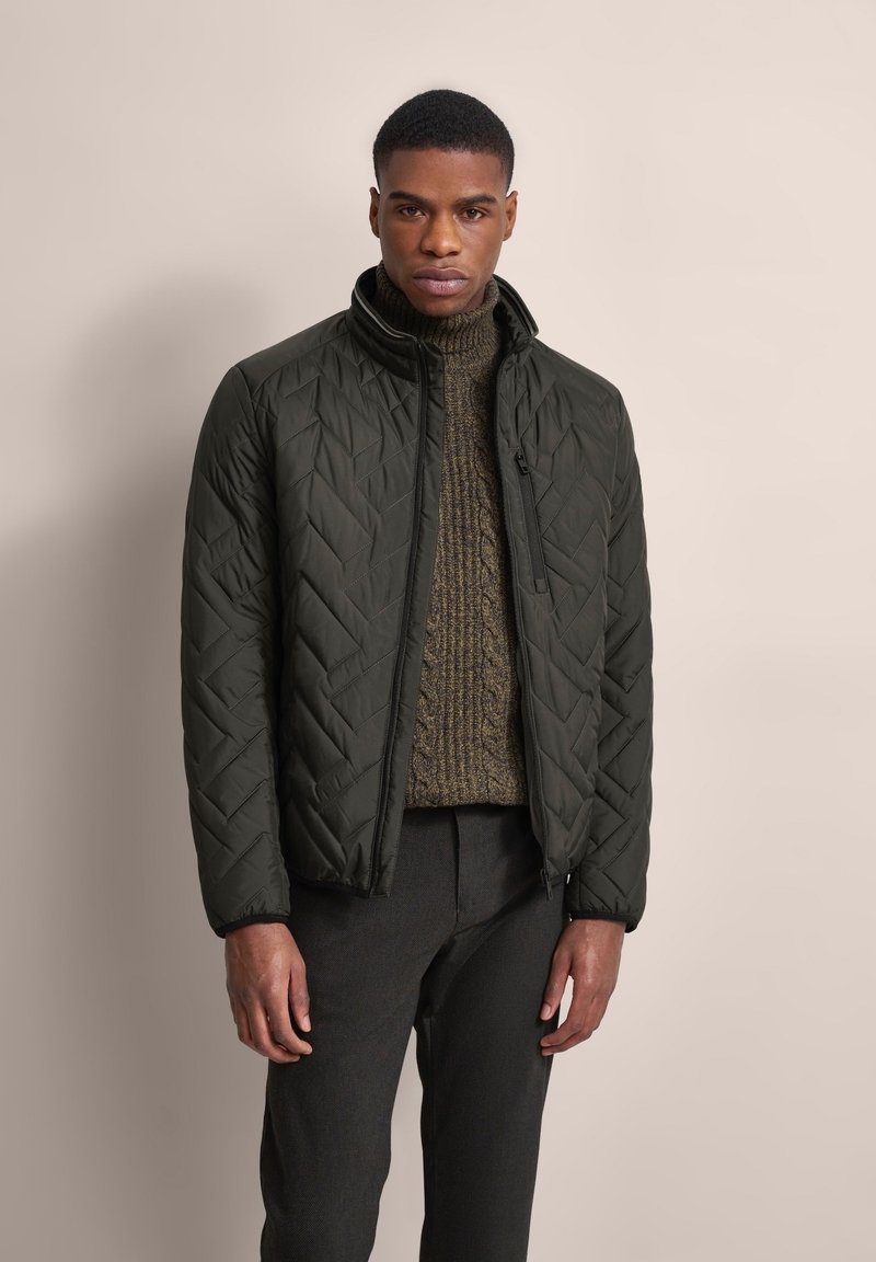 Dark green quilted jacket with a high collar and zip closure, worn over a textured brown sweater. Model poses against a neutral background.