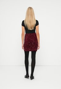 Bordo mini suknja s glitterom koja slijedi tijelo, u kombinaciji s crnom kratkom majicom s kratkim rukavima i crnim tajicama, prikazana sa stražnje strane.