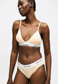 Μαύρη γυναίκα φοράει μπεζ bralette Calvin Klein και ταιριαστό εσώρουχο, στέκεται μπροστά σε απλό λευκό φόντο.