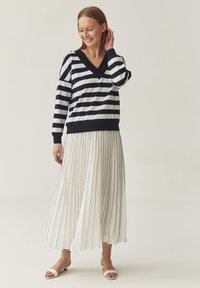 Maglione a righe blu navy e bianche con scollo a V e maniche lunghe abbinato a una gonna midi bianca plissettata con strisce verticali blu. Sandali piatti bianchi.