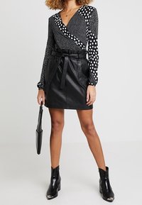 Czarna skórzana mini spódnica z wysokim pasem i wiązanym paskiem. Bluzka ma mieszane wzory czarnych i białych kropek, długie rękawy oraz dekolt w kształcie litery V.