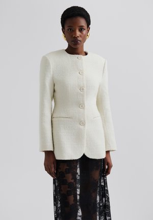 White textured blazer with a round neckline, five buttons, and two front pockets, worn over a black lace skirt.