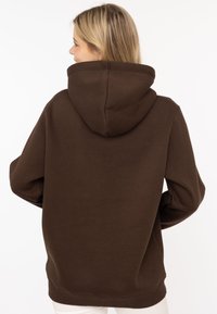 Brązowa bluza z dużym kapturem, z opadającymi ramionami i ściągaczami przy mankietach. Miękka tkanina, luźny krój i boczne kieszenie niewidoczne z tyłu.