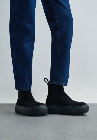 Noge u tamnoplavim farmerkama i crnim cipelicama bez pertli s debelim đonom, stoje na sivoj podlozi uz svijetlu pozadinu.