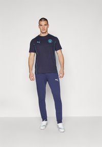 Tumši zili sporta T-kreklis ar gaiši ziliem akcentiem un logotipu, apvienots ar tumši ziliem sporta biksēm. Abi izgatavoti no viegla auduma komfortam.