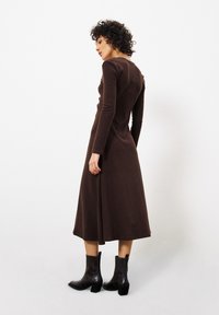 Braunes Langarmkleid, am Oberteil tailliert, mit weitem Saum. Strukturiertes Gewebe mit glatter Oberfläche. Getragen mit schwarzen Knöchelstiefeln.