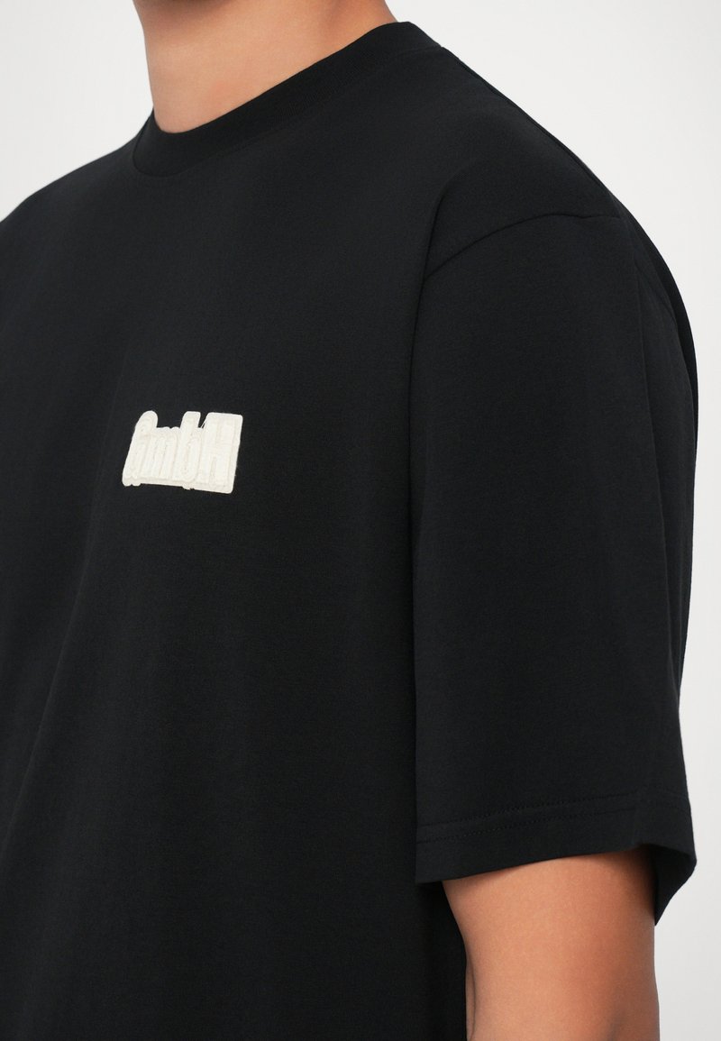 Sort bomulds t-shirt med rund halsudskæring og korte ærmer. Har et hvidt broderet "gmbH" logo på venstre bryst. Glat tekstur.