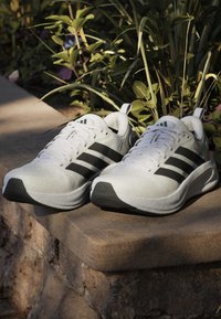 Paire de chaussures de course blanches Adidas avec des bandes noires posées sur un rebord en pierre à l'extérieur près de plantes vertes.