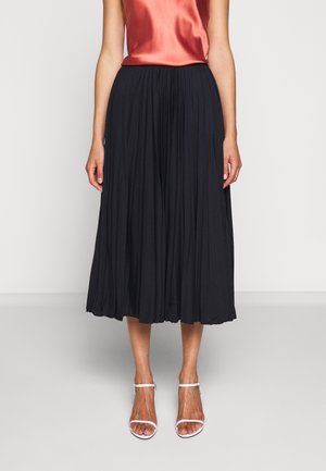 Femme portant une jupe midi plissée noire, un top sans manches en satin corail et des sandales blanches à talons ouverts, debout devant un fond uni.