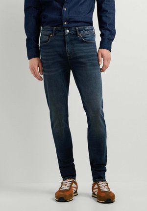Hombre vestido con vaqueros de mezclilla azul oscuro, zapatillas marrones y blancas, y una camisa azul oscuro abotonada, de pie frente a un fondo liso.