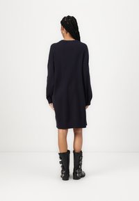 Vestido de suéter azul marinho com mangas longas, punhos canelados e fendas laterais. Usado com botas pretas até o joelho com fivelas prateadas.