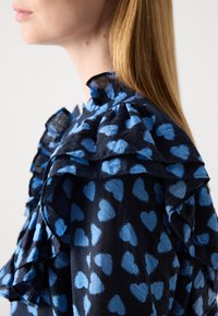 Blusa navy con colletto arricciato, caratterizzata da un motivo di cuori blu. Realizzata in tessuto leggero con finitura testurizzata.