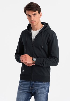Black zip-up hoodie with a drawstring hood, ribbed cuffs, and hem. Features a front pocket and a small logo patch at the bottom.