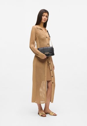 Ženska, oblečena v dolg bež kardigan iz mrežastega pletenja čez rjavo obleko, drži črno torbico in nosi rjave natikače.