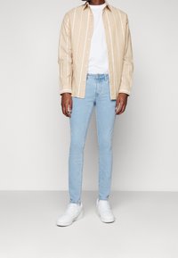Hombre con camisa de rayas verticales beige sobre camiseta blanca, jeans ajustados de color azul claro y zapatillas blancas, de pie sobre un fondo neutro.