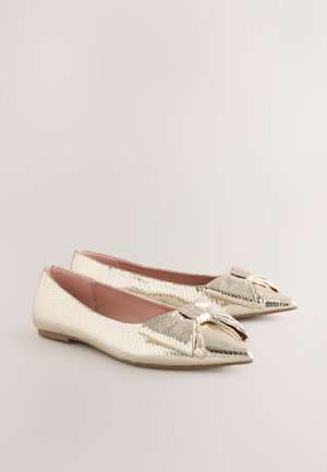 Scarpe basse dorate con un motivo testurizzato a squame, punta appuntita e fiocco decorativo sulla parte superiore. La fodera interna è rosa e la suola è piatta.