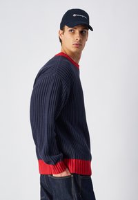 Pull en maille côtelée marine avec des poignets rouges, doté d'un col rond. Porté avec un jean en denim sombre. Une casquette marine avec le logo "Champion" est également visible.
