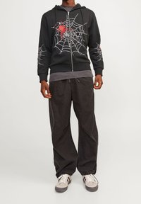 Sudadera negra con cremallera, que presenta un gráfico de telaraña blanca y una araña roja en la parte frontal y en la manga. Combinada con pantalones holgados de color gris oscuro y zapatillas ligeras.