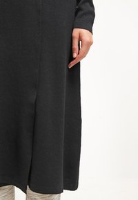 Persona che indossa un lungo vestito nero con uno spacco laterale e leggings chiari a fantasia, mano rilassata accanto al vestito.