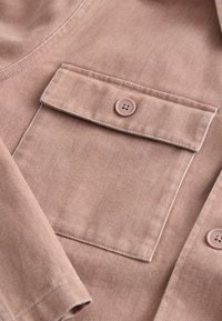 Gros plan sur la poche d'une veste en tissu marron clair avec rabat et bouton unique, montrant des coutures fines et un matériau texturé.