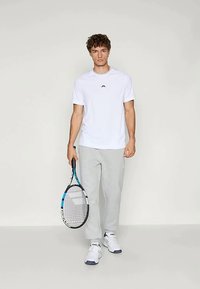 Bílý tričko s krátkým rukávem a malým logem, spárované se světle šedými tepláky, držící tenisovou raketu, obuté v bílých atletických botách.