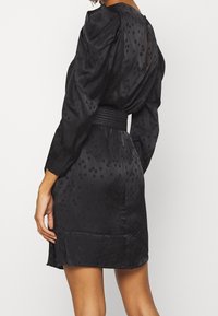 Černé šaty s texturovaným, tečkovaným vzorem, dlouhými rukávy a tkaným páskem v pase. Jemně rozšířený lem. Hladká látka.