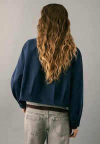 Chaqueta azul marino corta con mangas holgadas, textura suave, un solo botón en el puño, usada sobre un suéter gris y jeans gris claro.