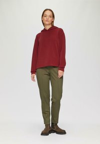 Bordeauxrotem Hoodie mit Kordeln, lockere Passform, kombiniert mit olivgrünen, schmal zulaufenden Hosen. Das Model trägt braune Stiefeletten. Einfacher Hintergrund.