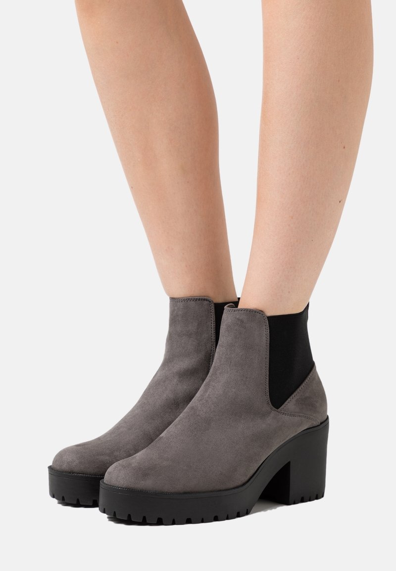 Bottes en daim gris montant à la cheville avec talons compensés noirs épais portées sur des jambes nues sur un fond uni.