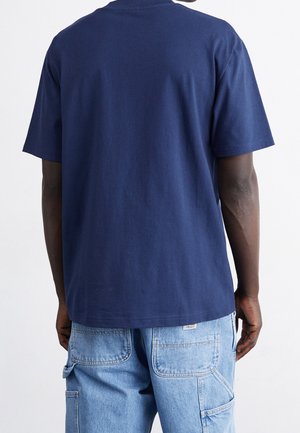 Person iført en ensfarvet mørkeblå kortærmet T-shirt og lyseblå denim cargobukser, set bagfra.
