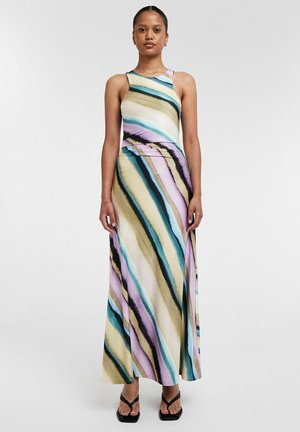 Vrouw draagt een mouwloze jurk tot op de vloer met diagonale pastelkleurige strepen in turquoise, paars, beige en zwart, gecombineerd met zwarte sandalen.