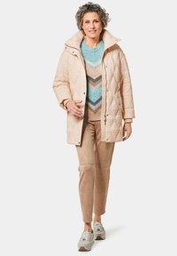 Beige gesteppte Jacke mit hohem Kragen, getragen über einem mehrfarbigen Pulli mit Zickzackmustern, kombiniert mit hellbraunen Hosen und Sneakers.