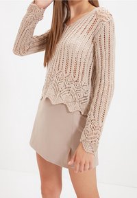 Beżowy sweter dzianinowy z długimi rękawami, z teksturowanym wzorem i falbaniastym dołem, noszony na beżowej spódnicy z bocznym rozcięciem.
