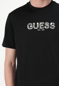 Juodas medvilninis marškinėliai su balta "GUESS" logotipu ir spauda "EST. 1981". Trumpomis rankovėmis ir apvaliu iškirpimu.