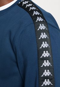 Épaule d’un vêtement bleu avec une bande noire présentant un logo blanc répété de deux silhouettes assises dos à dos.