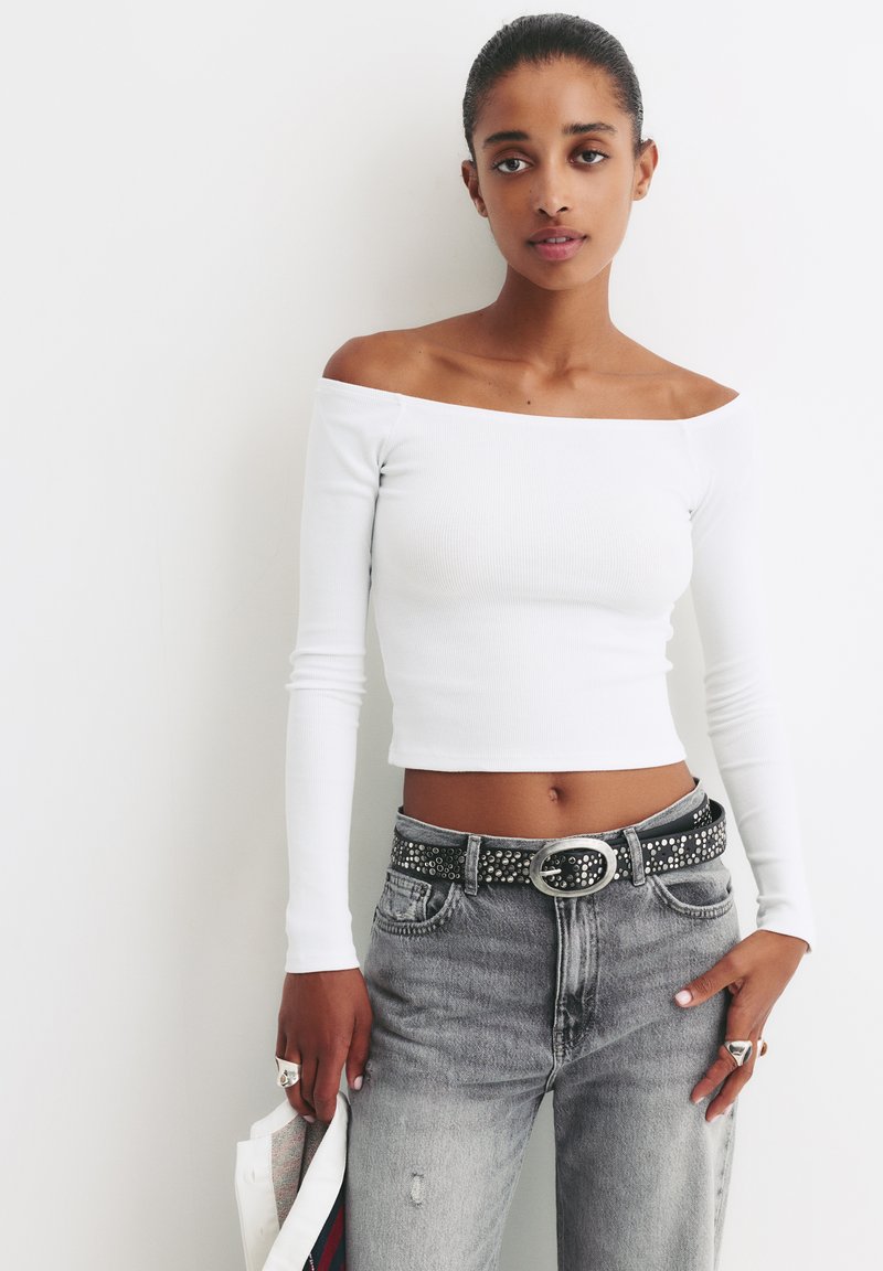 Bílý crop top bez ramínek se žebrováním, v kombinaci se šedými džínami s vysokým pasem a černým opaskem s cvočky, prezentující uvolněný, moderní střih.