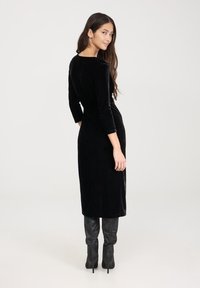 Czarna aksamitna sukienka midi z długimi rękawami i konturowaniem po bokach. Gładka tekstura, dopasowany krój, w połączeniu z czarnymi kozaczkami na obcasie.